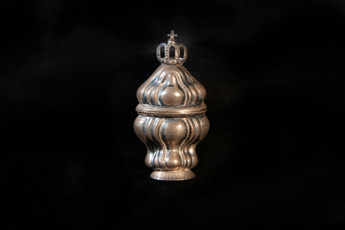 Hög dosa av silver i rokoko. Dekorerad med så kallade "knäckta åsar" i relief, och högst upp på locket en genombruten krona med kors. Utländska stämplar i botten. Troligen dansk tillverkning. Jämför med JM 18 742 och 19 952.