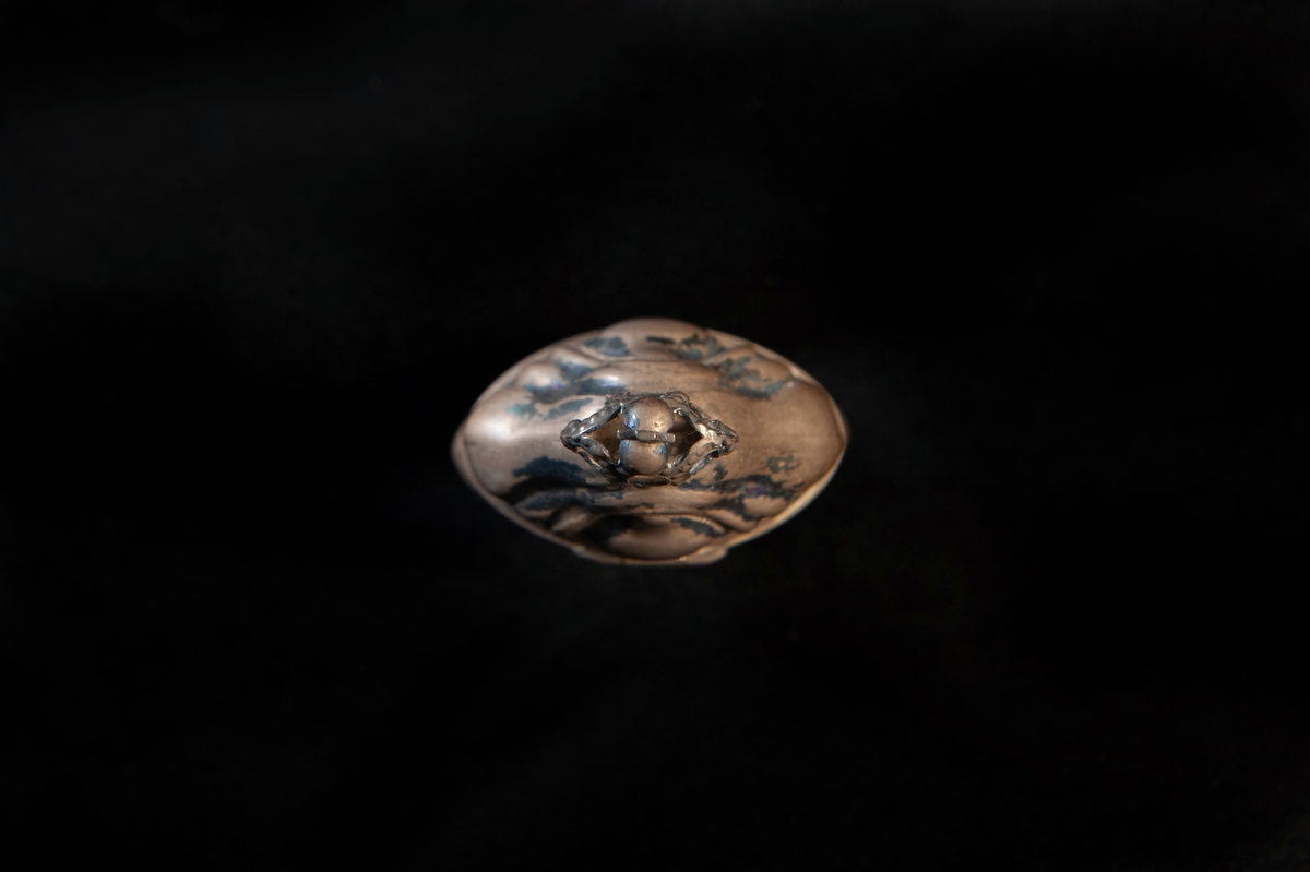 Hög dosa av silver i rokoko. Dekorerad med så kallade "knäckta åsar" i relief, och högst upp på locket en genombruten krona med kors. Utländska stämplar i botten. Troligen dansk tillverkning. Jämför med JM 18 742 och 19 952.