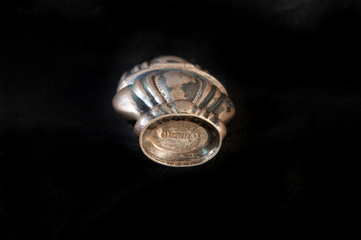 Hög dosa av silver i rokoko. Dekorerad med så kallade "knäckta åsar" i relief, och högst upp på locket en genombruten krona med kors. Utländska stämplar i botten. Troligen dansk tillverkning. Jämför med JM 18 742 och 19 952.