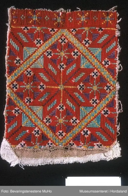 Rektangulær bringeduk med "enkel" og dobbel kross-sting i forskjellige retningar. Stor åttebladrose i midten og fire små oppe, nede og på sidene med ei rame rundt. I kvart hjørne er det eit blad av åttebladrose. Bord oppe med anna mønster (slags stjernemønster).  Raud hovudfarge. Dei mørkeblå krossane er sydd med kross-sting. Fargane er litt falma, spesielt den grønturkise og gule.