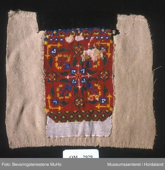 Rektangulært broderi på kvadratisk bakstykke. Plattsaum (halve kross-sting) i fleire retningar sydd på stramei. Nokre usydde og utslitte område. Raud hovudfarge. Diverse geometriske figurar og ei slags åttebladrose i midten. Bord nede  og oppe. Broderistoffet går litt nedom den nederste borda. Svart kant oppe. Alt er sydd på eit ullstoff vove i kypert. Fleire hol.