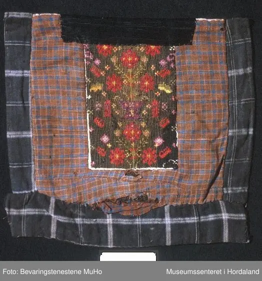 Rektangulær brystduk på kvadratisk botnstykke. Brystduken er brodert med halve kross-sting på stramei. Blomstermotiv i mange skarpe fargar. Ein svart silkeband oppe. Bomullstoff til botnstoff, bakstykke og sidestykke og stykke nederst. To forskjellige stoff til botn stoff med ruter. Brystduken er forsterka inni med tjukk kypertstoff.