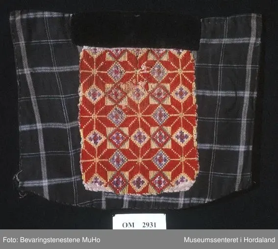 Rektangulær brystduk som er brodert i kross-sting (halve og heile). Åtteblad-rose som hovudmotiv og ruter. Broderiet er litt slitt. Svart fløyelsband oppe. Broderiet er sydd på linstoff. Bakstykket og sidestykke er i bomull - rutete mønster.