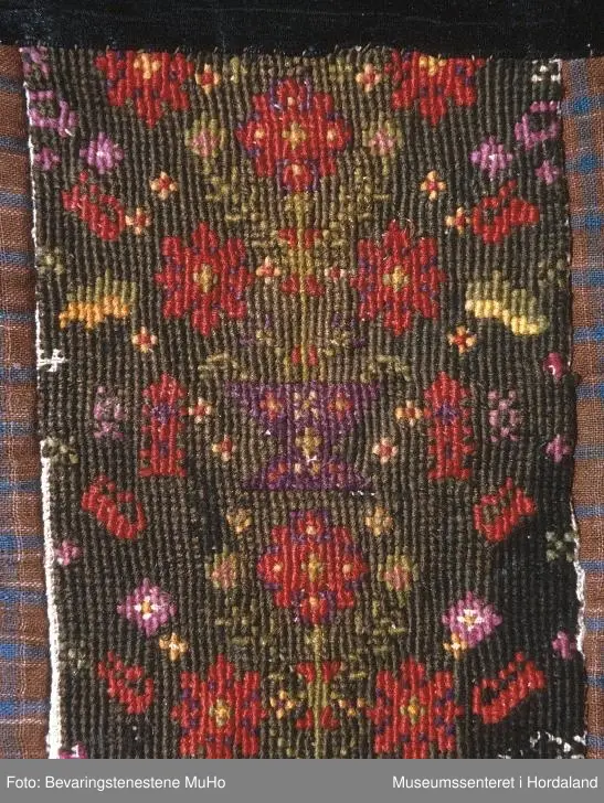 Rektangulær brystduk på kvadratisk botnstykke. Brystduken er brodert med halve kross-sting på stramei. Blomstermotiv i mange skarpe fargar. Ein svart silkeband oppe. Bomullstoff til botnstoff, bakstykke og sidestykke og stykke nederst. To forskjellige stoff til botn stoff med ruter. Brystduken er forsterka inni med tjukk kypertstoff.