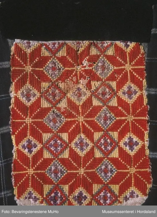 Rektangulær brystduk som er brodert i kross-sting (halve og heile). Åtteblad-rose som hovudmotiv og ruter. Broderiet er litt slitt. Svart fløyelsband oppe. Broderiet er sydd på linstoff. Bakstykket og sidestykke er i bomull - rutete mønster.