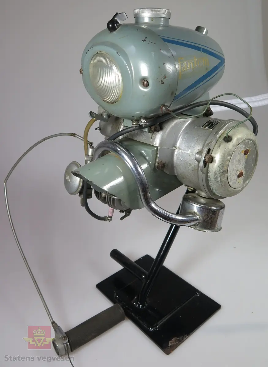 Fantom Bj.52. 48 ccm forbrenningsmotor med tank og lys. Er her montert på et stativ. Er lagd for å kunne monteres foran på sykkel slik at gummi hjulet på denne motoren skal dra rundt hjulet på sykkelen. Motoren har 1,4 hk og er av typen 4-takt, den går på ren bensin.