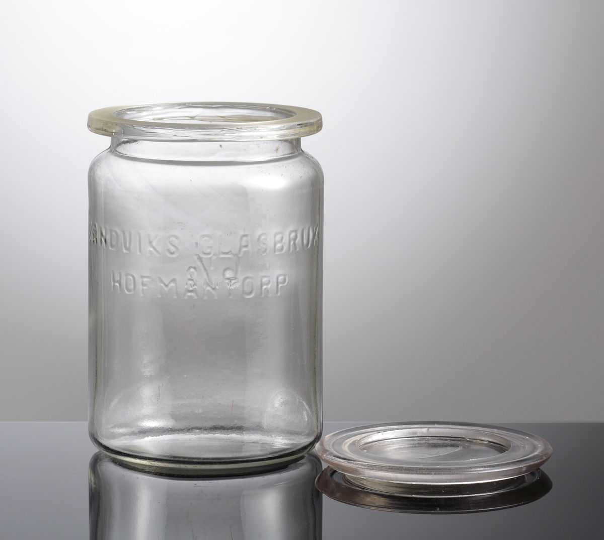 Konserveringsburk med lock.
Tillverkad på Sandviks glasbruk.
Cylindrisk, kraftig krage, planslipad matt mynning.
Upphöjd relief med text: "Sandviks glasbruk Hofmantorp"
Blåst i tvådelad form. Locket pressat.