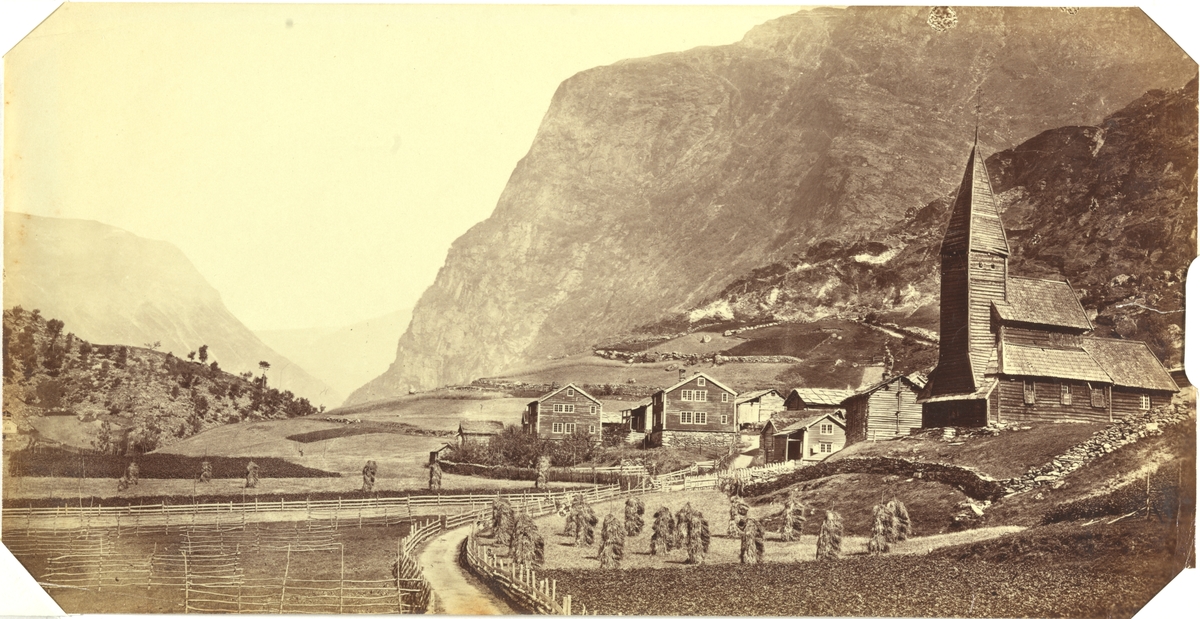Fortun stavkirke (revet 1883), Luster i Sogn. Ved kirken ligger tømmerhus, på markene står kornstakker. Stein- og tregjerder deler inn området. Fjell i bakgrunnen. Antagelig fotografert ca. 1873.