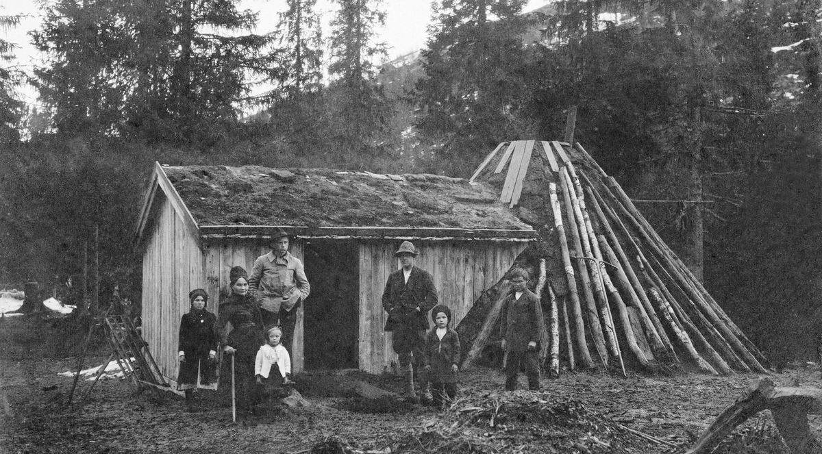 Familiegruppe ved gamme ved Gramfjell i Vefsn.  Gamma framstår som en kjegleformet konstruksjon, dekt med torv, bjørkerajer og bord.  Inntil gamma er det bygd et rektangulært, bordkledd skur med torvtak.  Foran bygningene står to menn, ei kvinne og fire barn.  Flere av dem har drakter som tyder på samisk bakgrunn. 
