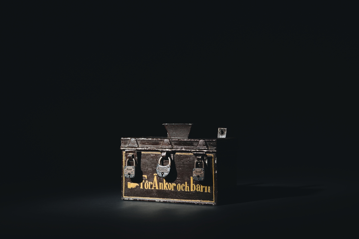 Sparbössa rektangulär, på översidan försedd med lock och öppning för pengarna, tre rörliga överfall för låsning med tre stycken hänglås.