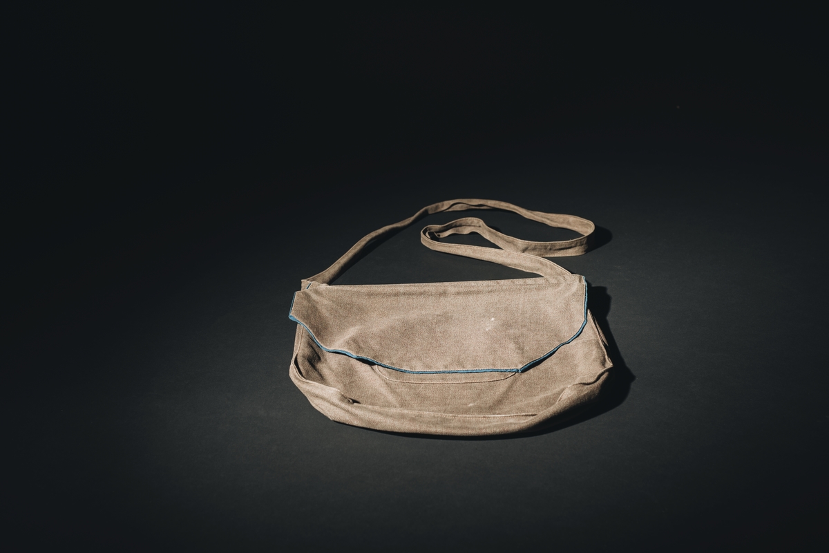 Axelväska för marinlotta i gråbrungrönt tyg. Sydd som en påse med bälg. Lock, som täcker halva framsidan med mellanblått fodertyg som också bildar passpoal runt lockets framsida. Under locket dragkedjsöppning till väskpåsen samt en mindre utanpåsydd ficka också med dragkedjsöppning. Bakstycket slät. I ovankantens hörn är axelrem fästad.  Märkt under locket: W, med blå tråd. I väskan upphittades text av lottasång.
