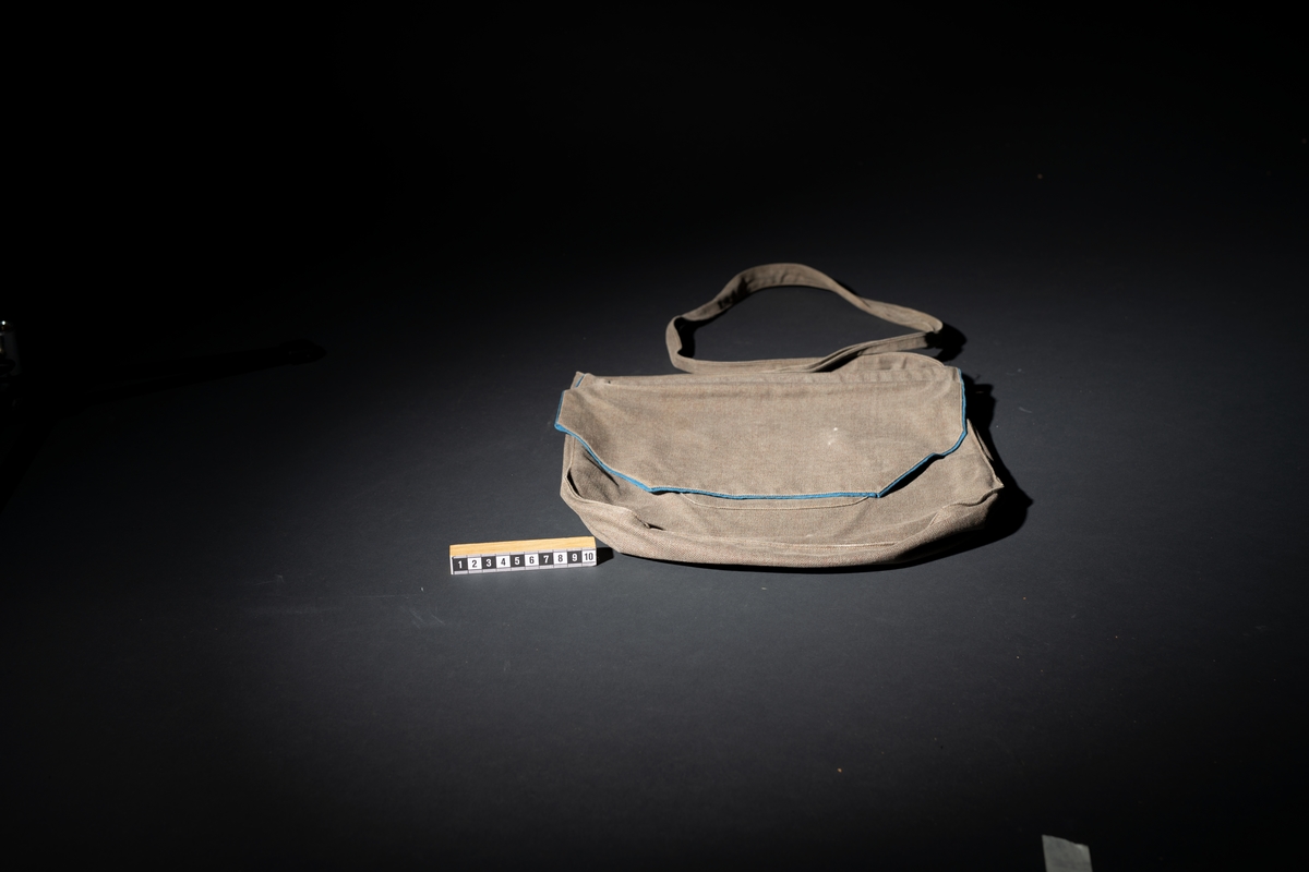 Axelväska för marinlotta i gråbrungrönt tyg. Sydd som en påse med bälg. Lock, som täcker halva framsidan med mellanblått fodertyg som också bildar passpoal runt lockets framsida. Under locket dragkedjsöppning till väskpåsen samt en mindre utanpåsydd ficka också med dragkedjsöppning. Bakstycket slät. I ovankantens hörn är axelrem fästad.  Märkt under locket: W, med blå tråd. I väskan upphittades text av lottasång.