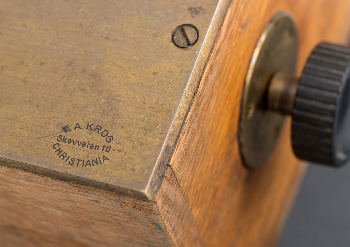 Kubikkberegningsapparat, levert av produsenten F. A. Krog i Skovveien i Kristiania, antakelig tidlig på 1900-tallet.  Apparatet er formet som et rektangulært skrin, utført i eiketre, men med "lokk" av ei messingplate.  Langs midtaksen på dette lokket er det ei lang, smal, rektanuglær spalte med glass under.  Gjennom denne ser vi ned på en tabell.  Tabellen er trykket på en papirrull, som er forankret på to valser nede i skrinet.  Tabellen kan beveges ved hjelp av dreiehjul på skrinets endeflater, to på hver side, ett i hver ende av de nevnte valsene.  Lengst til høyre i tabellvinduet finner vi "klassifiseringsinformasjon" - angivelser av stokklengder, diametere og salgsklasser.  Langs den øvrige delen av vinduet kan man lese av kubikkmeterangivelser for fra 1 til 70 stokker med samme dimensjoner og klassespesifikasjon.  Avlesinga skjer fra en tolinjet tabell på den nevnte papirrullen inne i skrinet, og med stokketallesangivelser på lokket - 1-35 stokker på oversida av vinduet og 36-70 stokker på undersida.  Det nevnte emaljerte lokket, som er hvitt med svarte tall og bokstaver, er skrudd fast til toppen av skrinet.  Det er grunn til å tro at valsene med tabellen må være montert før dette ble påsatt.  Skrinet er ellers lagd av høvlet eiketre, og den holdes sammen av de nevnte skruene i lokket og spiker fra botnstykket.  På standflata er det også hengslet en klaff, som kan vippes opp, slik at lokket skrår mot brukeren når apparatet brukes på et bord eller et annet plant underlag. 