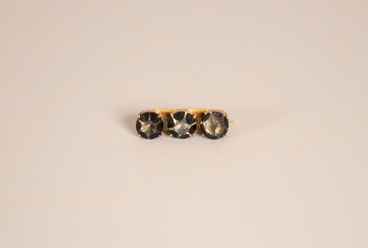 Langstrakt, plate med tre store stener i dyp innfatning. Stenene fasettslipt, den ene har en skarp spiss de to andre stenene er flate på toppen. Sortlakkerte tunger langs sidene, malt i ulike lengder så steinene får hvert sitt uttrykk. Hengslet  festenål.