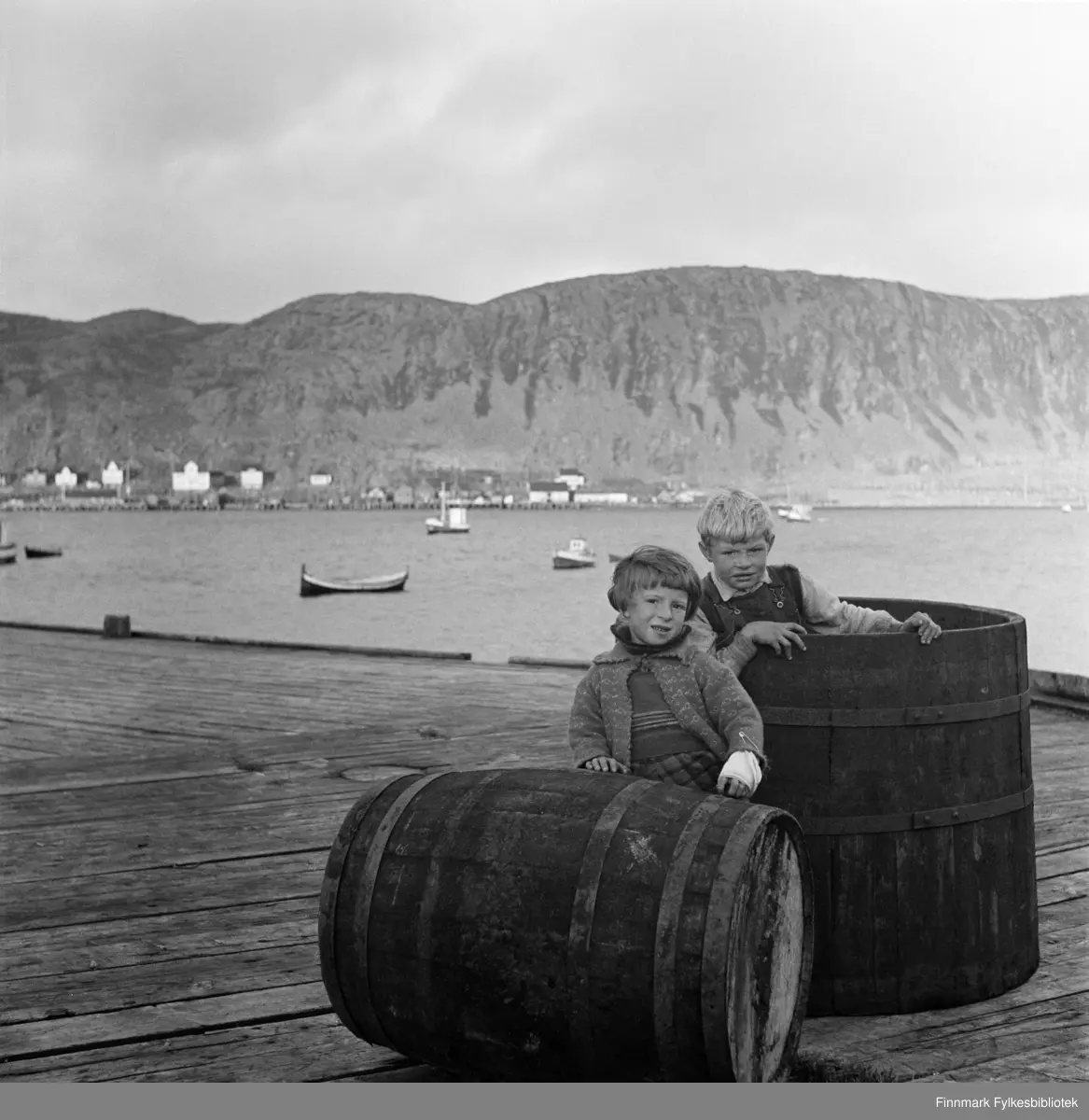 Åse Isaksen og Alf Nilsen (født 15.12.1933) på Nissen-kaia i Kjøllefjord, fotografert i 1940. Åse har bandasje på venstre arm med stor sikkerhetsnål. På andre siden av bukta sees flere fiskebruk og fiskemottak, hotell og boliger i Kjøllefjord. På fjorden ligger fiskebåter oppankret. En nordlandsbåt nærmest kamera. Åse har en strikket jakke og genser på seg og rutete skjørt eller bukser. Alf ser ut til å ha på seg snekkerbukser og skjorte. Sannsynligvis er dette på Nissenkaia.
