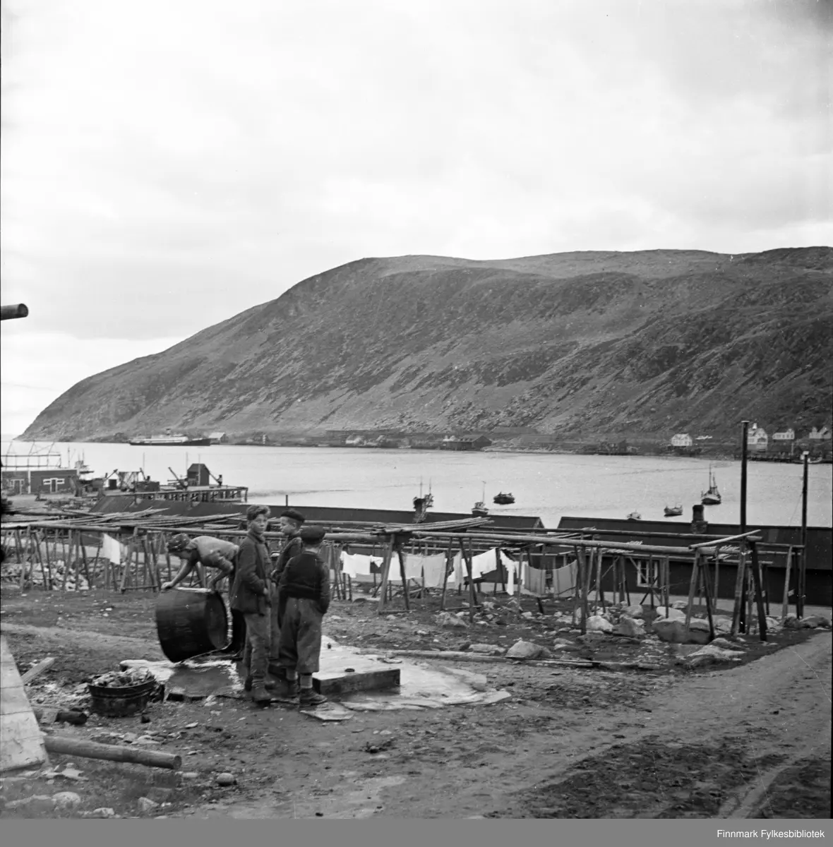 Kjøllefjord 1940. Tre gutter står og henger, mens en ung jente tømmer stampen etter klesvasken som henger på snora i bakgrunnen. Klessnora er rett og slett hengt opp mellom fiskehjellene hvor tørrfisken henger på vinteren. I bakgrunnen ligger fiskebruk og flere fiskebåter ligger fortøyd utenfor. I bakgrunnen kan man se at Hurtigruteskipet DS «Prinsesse Ragnhild» ankommer Kjøllefjord og er i ferd med å legge til kai.