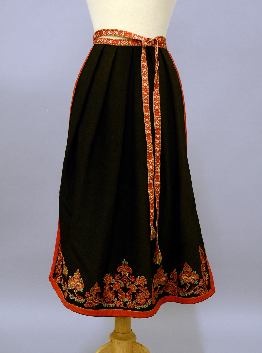 Modellbunad fra Vest-Telemark.TGM-BM.1999:0143.A-D.
B. Forkle. Sort forkle i ull med brodert bord i rosesøm nederst. Forkleet er kantet med et rødt og et grønt ullbånd. Påmontert forklebånd i grindvevteknikk i fargene rødt, hvitt, beige og grønt.