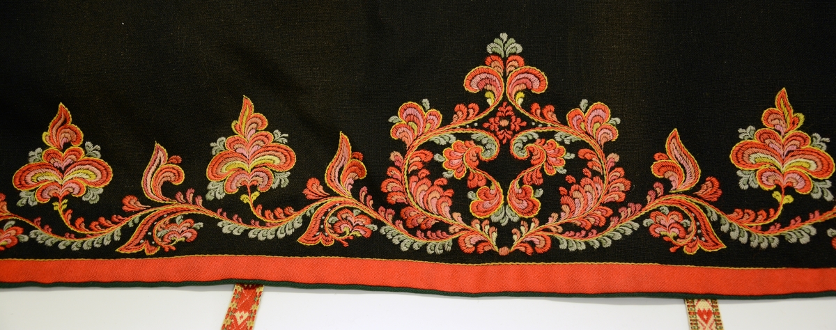 Modellbunad fra Vest-Telemark.TGM-BM.1999:0143.A-D.
B. Forkle. Sort forkle i ull med brodert bord i rosesøm nederst. Forkleet er kantet med et rødt og et grønt ullbånd. Påmontert forklebånd i grindvevteknikk i fargene rødt, hvitt, beige og grønt.