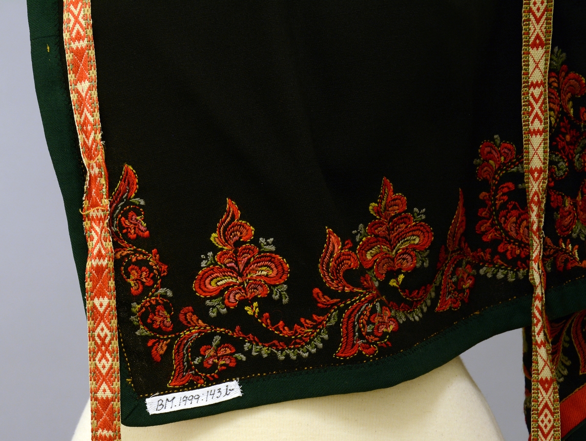 Modellbunad fra Vest-Telemark.TGM-BM.1999:0143.A-D.
B. Forkle. Sort forkle i ull med brodert bord i rosesøm nederst. Forkleet er kantet med et rødt og et grønt ullbånd. Påmontert forklebånd i grindvevteknikk i fargene rødt, hvitt, beige og grønt.