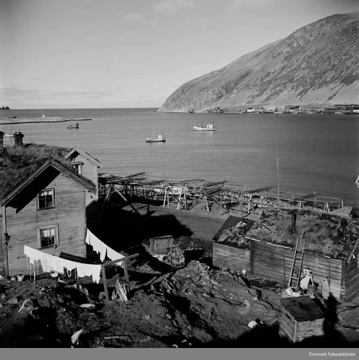 Klesvask til tørk utenfor huset. Kjøllefjord 1940. I bakgrunnen sees flere fiskebruk og fiskebåter ligger ankret i havna. To små jenter skygger for solen og titter i retning av fotografen. Bildet er tatt i nærheten av dagens LHL-brygge (Myrvoll?) iflg. vår informant.