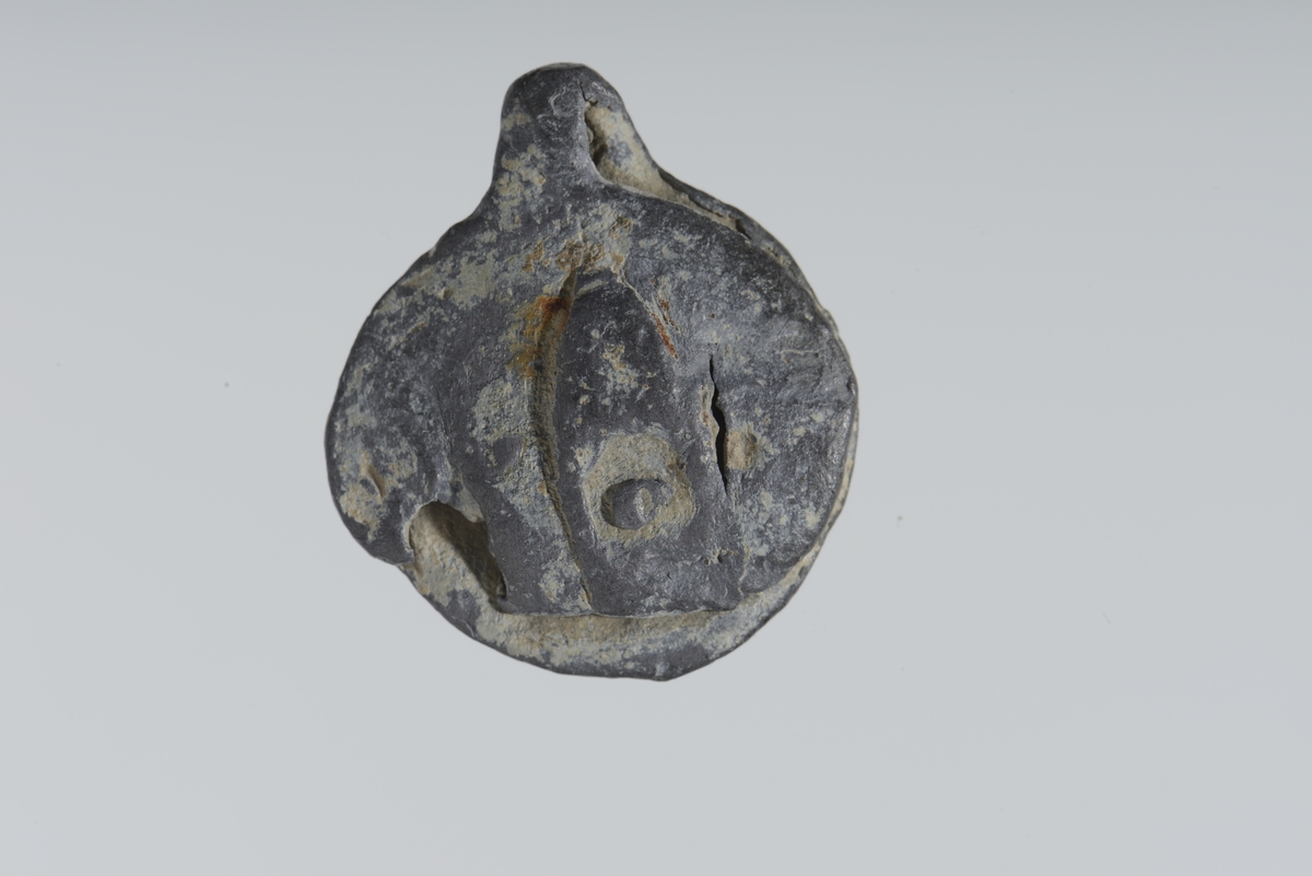 Vareplombe med begge plaketter, nagle og streng bevart. Gjenstanden er nedslitt men viser svake tegn til preg på begge plaketter. Preget er utydet. 