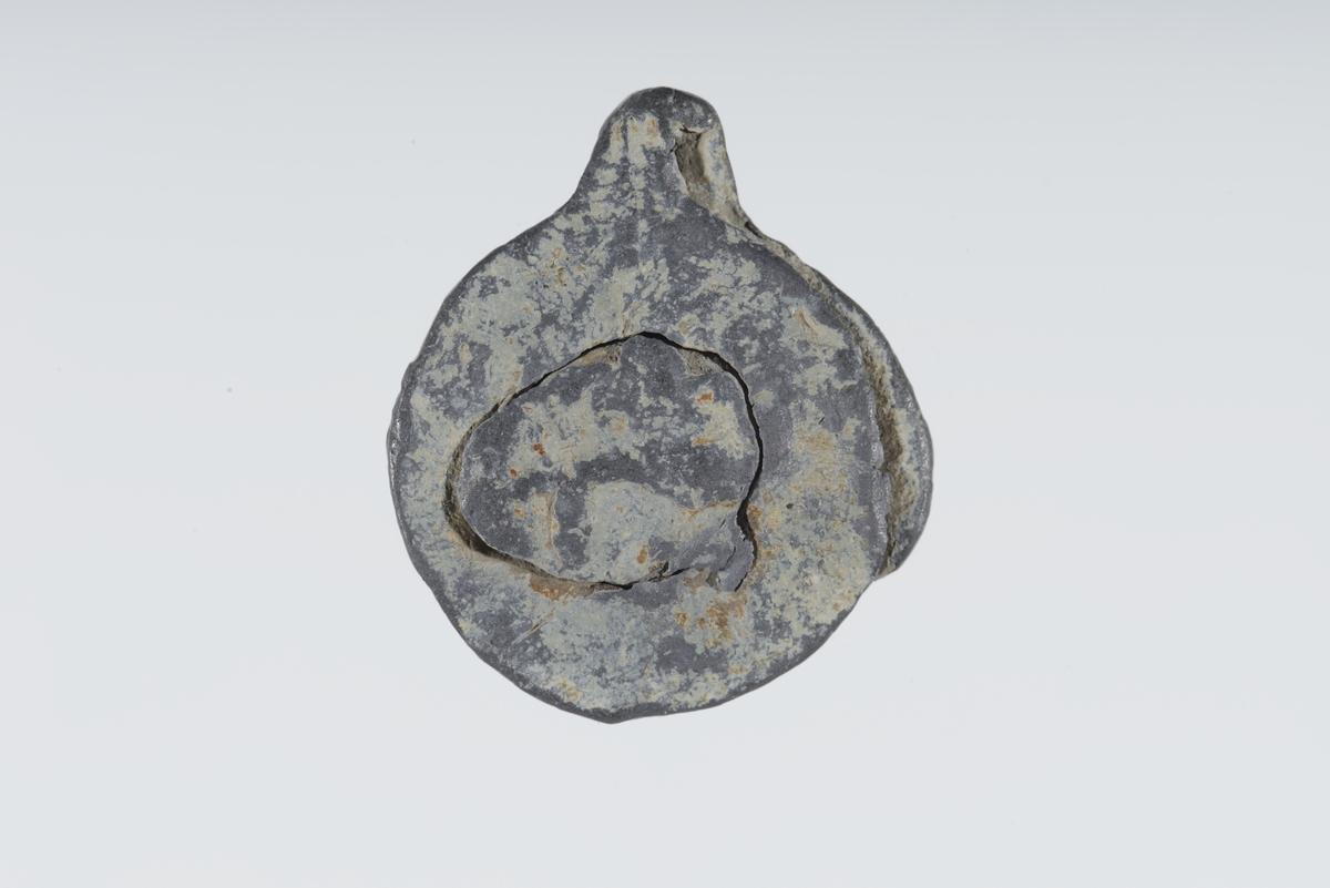 Vareplombe med begge plaketter, nagle og streng bevart. Gjenstanden er nedslitt men viser svake tegn til preg på begge plaketter. Preget er utydet. 