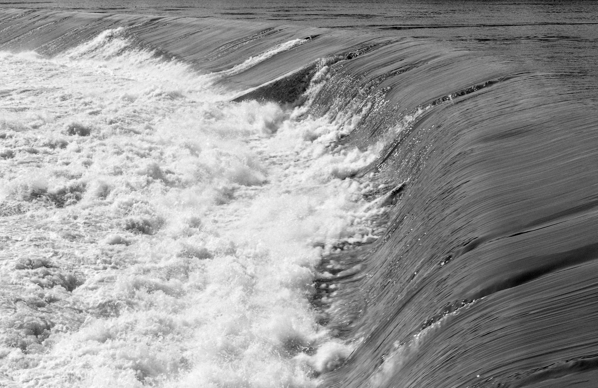 Fra kraftverksdammen øverst i Skjefstadfossen i Glomma i Heradsbygda i Elverum i Hedmark. Bildet ble tatt i 1961, fra en plattform ved det østre landsfestet, ned mot den 312 meter lange overløpsdammen, en buet betongkonstruksjon med den konvekse sida i motstrøms retning. Da dette fotografiet ble tatt gikk det ikke fløtingstømmer over damkrona, men øverst til høyre skimter vi lensa som skulle lede tømmeret mot den østre delen av elveløpet og fossen. Lensa var et flytende stengsel som skulle hindre at tømmeret drev inn i inntaksbassenget for vann til krafstasjonen, som lå lengre nede, ved vestre elvebredd. Mer informasjon om dammen og kraftverket ved Skjefstadfossen finnes under fanen «Opplysninger».