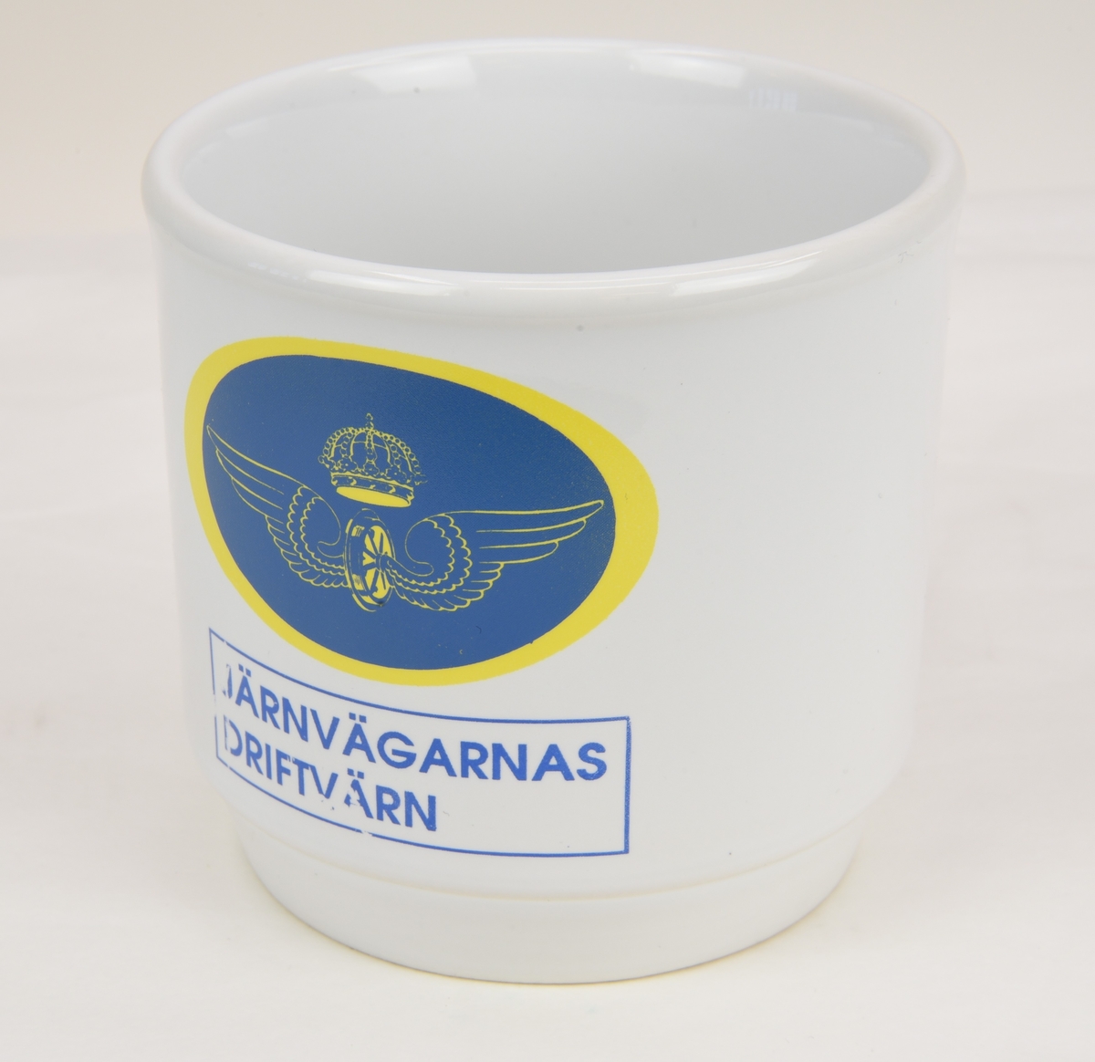 En vit kaffekopp av porslinmed tryckt motiv föreställande det bevingade samt krönta vinghjulet i en blå oval med gul kant. Nedanför texten "JÄRNVÄGARNAS DRIFTVÄRN" i en rektangel med en blå linje runtom.