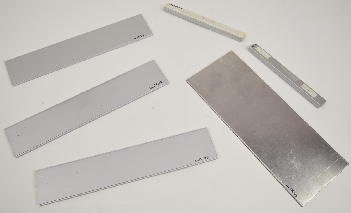 Namnskyltshållare av obehandlad extruderad aluminium (:1-3) med tillhörande väggfästen (:4-5) och en aluminiumplatta med vit yta som fungerar som en mini-whiteboard (:6). Skylthållarna har plats för två rader textremsor som skjuts in från sidan. En av hållarna saknar namn men två av dem har både namn och titlar med svart text på vita Dymo-remsor. Remsorna med "Teknikstöd" och "Fredrik Jansson" är inte fastklistrade utan har kvar Dymo-remsans täckpapper.