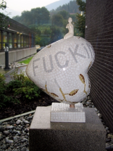 Liten hjerteformet skulptur. Armert gipskappe kledd med glassmosaikk. En liten porselensfigur er støpt/limt ned i skulpturen. Sokkelen er i rødlig granitt.
