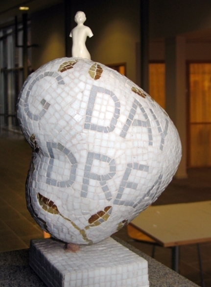 Liten hjerteformet skulptur. Armert gipskappe kledd med glassmosaikk. En liten porselensfigur er støpt/limt ned i skulpturen. Sokkelen er i rødlig granitt.