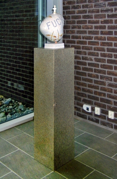 Liten hjerteformet skulptur. Armert gipskappe kledd med glassmosaikk. En liten porselensfigur er støpt/limt ned i skulpturen. Sokkelen er i rødlig granitt.
