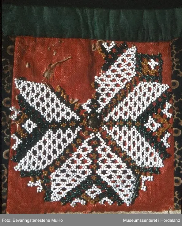 Rektangulær bringeduk med perlenett med åttebladrosemotiv sydd på raudt ullstoff. Perleveven er noko rakna og manglar eit blad. Midt i åttebladrosa er det sydd på ein knapp. Ullstoffet har fleire hol. Grøn silkeband oppe. Blått mønstrete bomullstoff på kvar side sydd på eit kypertvevd naturfarga stoff. Skittent på baksida. Det grøne bandet er sydd oppå eit anna rosafarga silkeband.