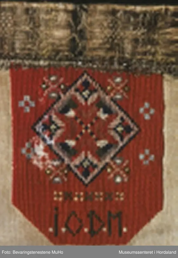 Rektangulær bringeduk sydd på kvadratisk bomullstoff. Broderiet er i halve og heile kross-sting sydd på linstoff. Raud hovudfarge. Initialane I O D M er sydd nederst på broderiet. Silkeband øvst med ei smal metallbord nedom. Silkebandet, metallborda og broderioet er veldig slitt.