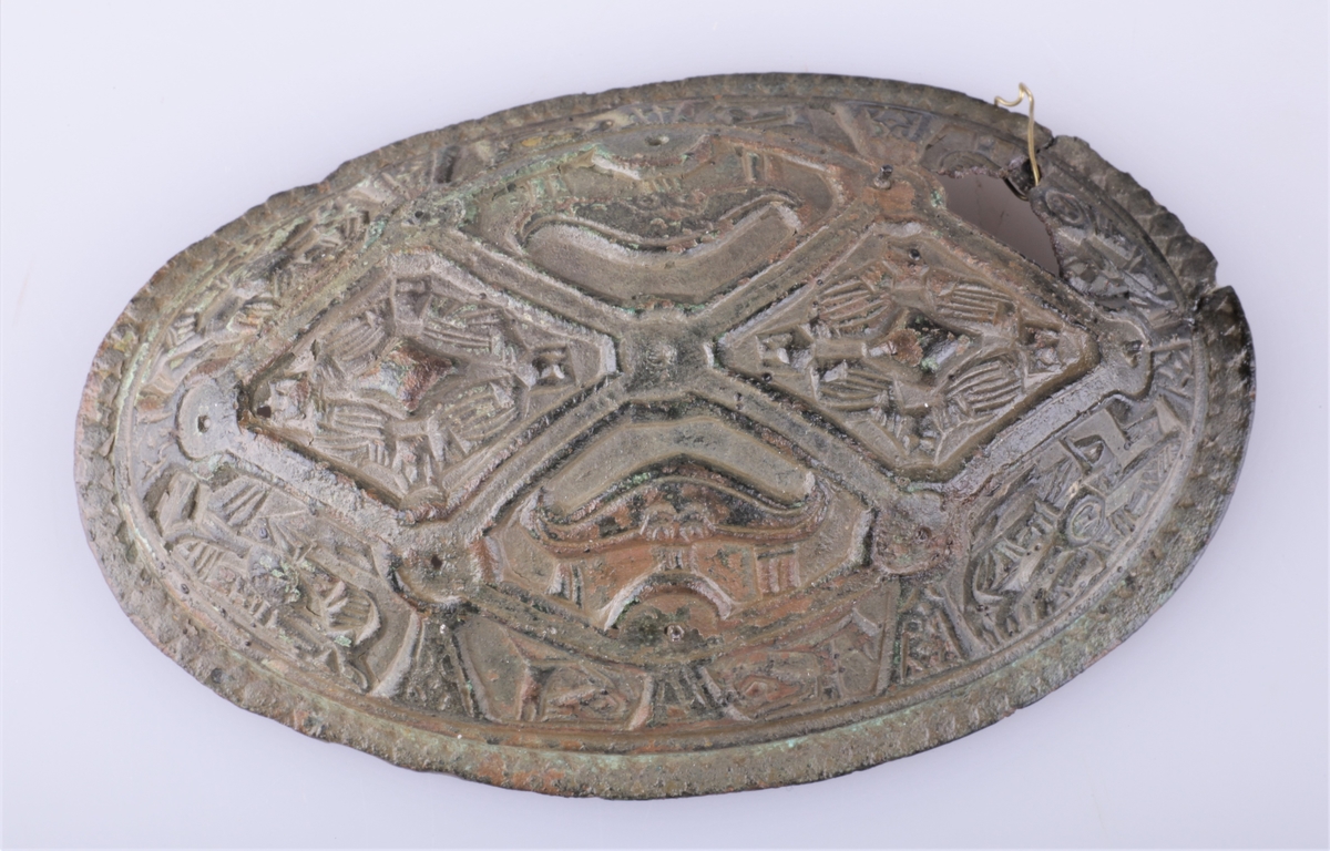 Oval bronsespenne med støpte ornamenter fra vikingtid. Variant av type Rygh 647. Funnet i en haug på gården Tråset i Fåberg i 1887.
