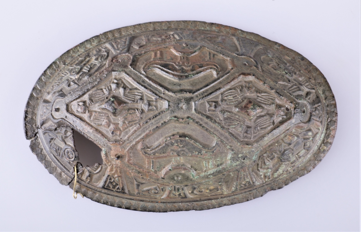 Oval bronsespenne med støpte ornamenter fra vikingtid. Variant av type Rygh 647. Funnet i en haug på gården Tråset i Fåberg i 1887.