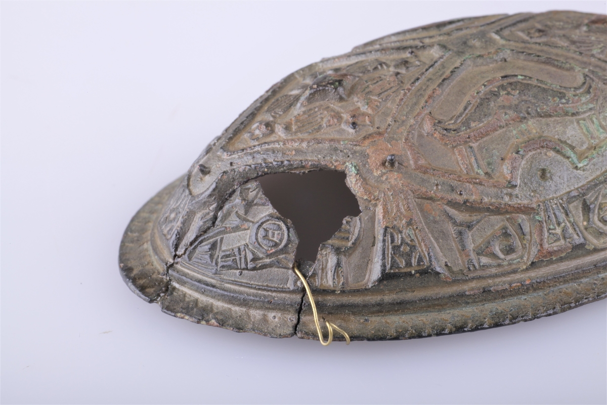 Oval bronsespenne med støpte ornamenter fra vikingtid. Variant av type Rygh 647. Funnet i en haug på gården Tråset i Fåberg i 1887.