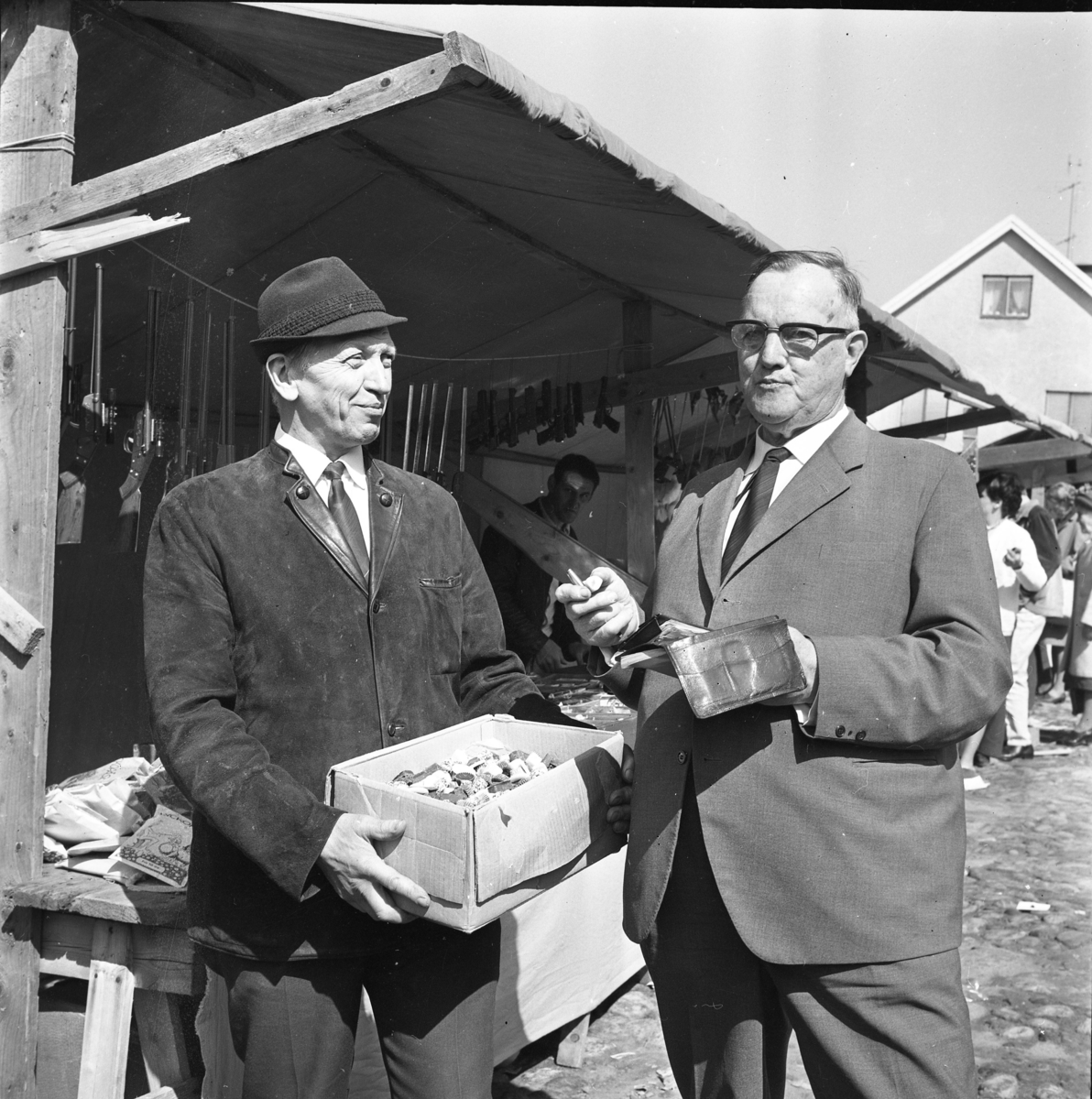 Höstmarknad i Gränna 1968. Gunnar Fransson - Gunnars-konfektyr - i hatt och mockajacka håller fram en kartong med karameller för marknadsföreståndaren Arvid Lindgren som låter sig smaka med plånboken uppslagen i näven. Det är möjligt att Lindgren, i kostym, går runt för att ta upp marknadsavgiften.