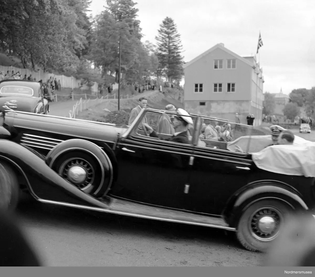Bilen er en Åpen Buick 1935 med kong Haakon VII i baksetet. (Info: 
Ivar Stav ). Foto fra 1000-årsjubileet for slaget på Rastarkalv på Frei i Kristiansund kommune. Slaget på Rastarkalv var et slag i 955 mellom Eirikssønnene og Håkon Adalsteinsfostre. Eirikssønnene ledet en hær som bestod av norske og danske vikinger, tildelt av den første danske kongen, Harald Blåtann. Håkon Adalsteinsfostre vant slaget og tvang danskene til retrett. (Kilde: Wikipedia). Bildet er datert 1955. Fotograf er Nils Williams. Fra Nordmøre museums fotosamlinger.