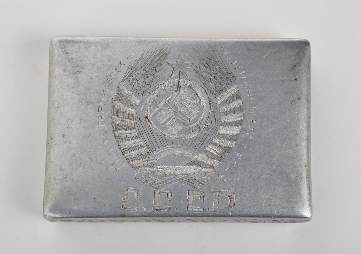 Sigarettetui laget av sovjetisk krigsfange. Etui tilvirket av aluminium med pregstempeldekor. På lokket er det risset inn ulike symboler, blant annet hammer, sigd og den røde stjerne, sammen med kornaks  og tekst. På eskens underside er arbeideren (med fane) avbildet sammen med tekst. Det har vært dekor både på lokkets innside og i bunne av etuit, men disse er blitt risset over.