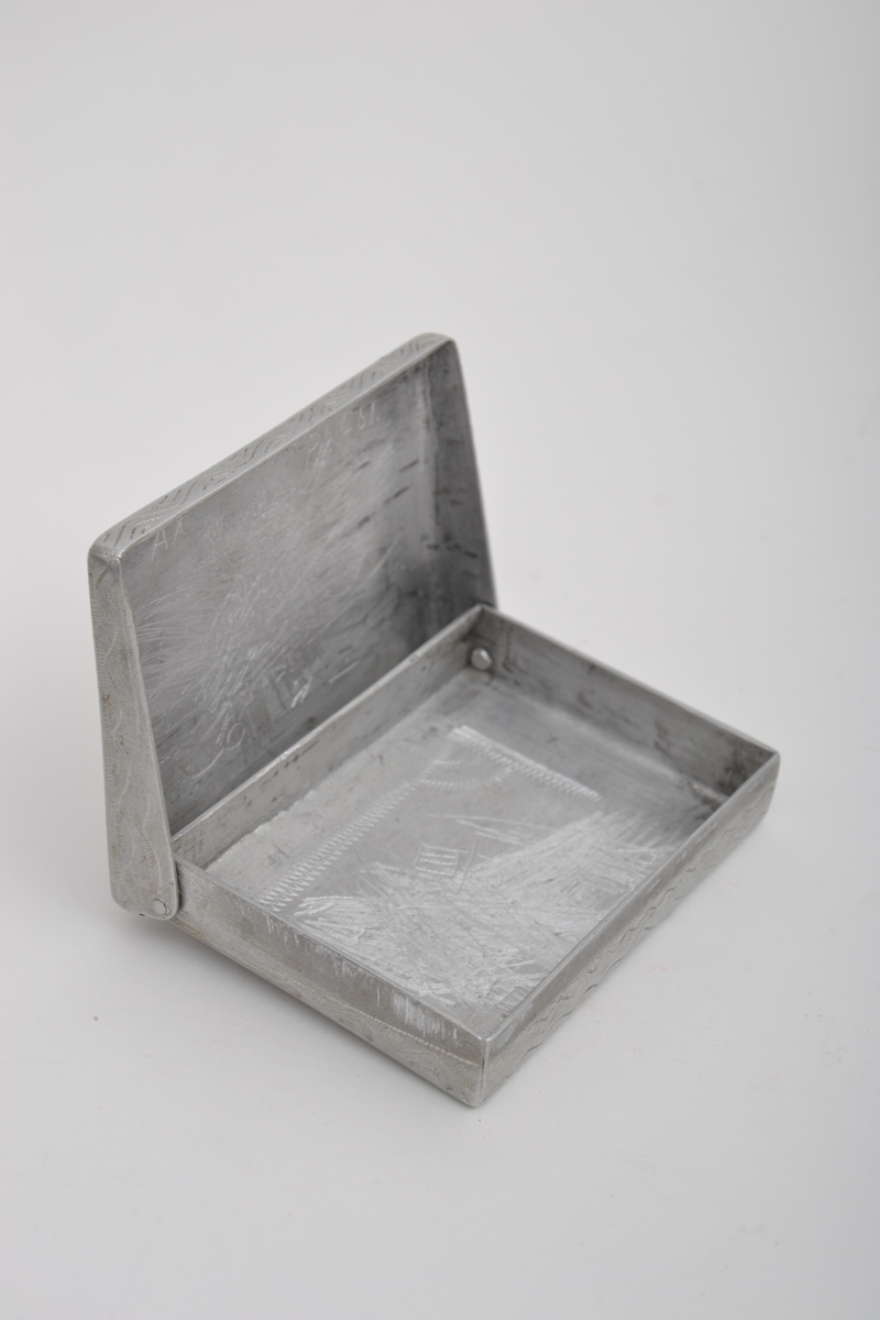 Sigarettetui laget av sovjetisk krigsfange. Etui tilvirket av aluminium med pregstempeldekor. På lokket er det risset inn ulike symboler, blant annet hammer, sigd og den røde stjerne, sammen med kornaks  og tekst. På eskens underside er arbeideren (med fane) avbildet sammen med tekst. Det har vært dekor både på lokkets innside og i bunne av etuit, men disse er blitt risset over.