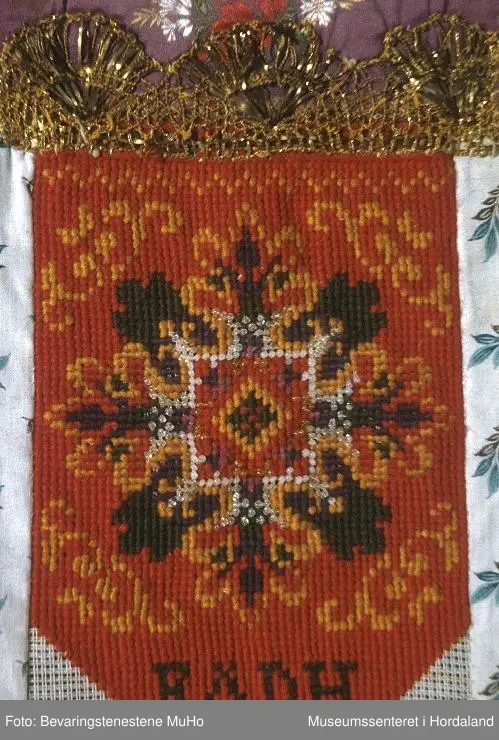 Bringeduk brodert med kross-sting på stramei i raudt, gult, grønt, lilla og kvite, blanke og gullfarga glasperler. Initialer er brodert nederst på broderiet. Blomstrete silkeband øvst. Kronekvarde i gull er montert eit stykke ned på broderiet og oppå silkebandet. Broderiet er montert på eit mønstrete bakstykke som og er brukt på kantane og under på framsida.