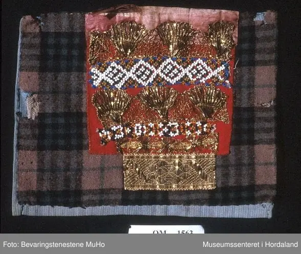 Bringeduk sett saman av ulike border. Øvst er montert ein rosa silkekvarde, under kjem ein kronekvare i gull, ei bord av perlenett, ein kronekvarde, ei smal bord av perlenett og ein smal kronekvarde, alt montert på raudt klede. Nedst, nedanfor kledet, ein liten bit av eit gullfarga metallband. Det raude kledet er montert på eit breiare, rutete ullstoff. Bakstykke av eit blått bomullsstoff.