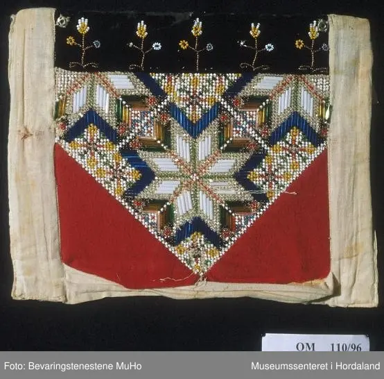 Perlebringekduk med åttebladroser som hovudmotiv. Her er brukt både små perler og ståperler. Perlene er sydde på stramei. Oppe er det ein svart pløyelskant der det er sydd på 5 stiliserte blomstermotiv i perler. Heilt øvst på fløyelstykket på kvar side er det sydd på trykk-knapper for å feste bringeduken til livet. Nedanfor perlene er det sydd på raude tøystykke i bomullsflanell. Utanfor sjølve perlemotivet er det kvitt lerret, også som bakstykke. Inni ligg det ei nokså stiv papp-plate. Bringkluten er velbrukt og slitt og manglar ein del perler.