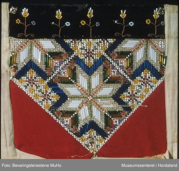 Perlebringekduk med åttebladroser som hovudmotiv. Her er brukt både små perler og ståperler. Perlene er sydde på stramei. Oppe er det ein svart pløyelskant der det er sydd på 5 stiliserte blomstermotiv i perler. Heilt øvst på fløyelstykket på kvar side er det sydd på trykk-knapper for å feste bringeduken til livet. Nedanfor perlene er det sydd på raude tøystykke i bomullsflanell. Utanfor sjølve perlemotivet er det kvitt lerret, også som bakstykke. Inni ligg det ei nokså stiv papp-plate. Bringkluten er velbrukt og slitt og manglar ein del perler.