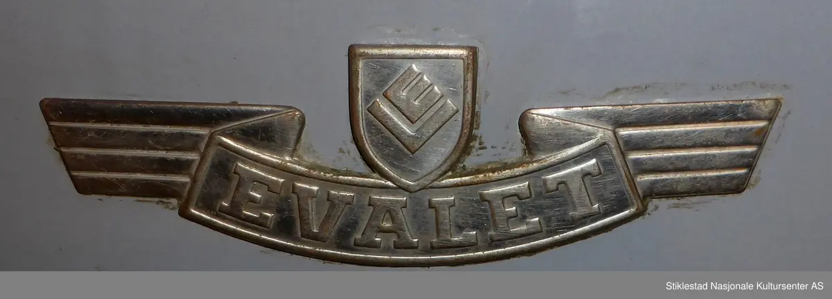 Elektrisk vaksemaskin i emaljert metall. Lokk i aluminium, gummislange morknet(knekt). Maskin med varmeelement som varmer opp vannet. Sluk i bunn av tank. Motor plassert under vanntank. Evalet logo i messing med vinger, plassert i front av maskin. Står på fire føtter, gummierte. Svart gummipakning i topp av maskin. Grep til lokk mangler. Består av 4 deler. Vaskemaskina hadde opprinnelig vrider som kunne monteres på toppen, denne kunne tas av og på. Denne har blitt kastet av giver (morknet)