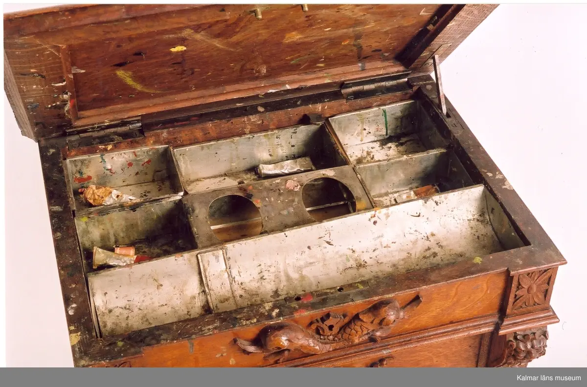 KLM 31928. Skåp. Av ek. Skåp med uppfällbar skiva och fyra utdragbara lådor. Utrymmet under skivan innehåller flera fack av förkromad plåt. I facken har funnits måleriutrustning, vilket bl.a. kan ses genom talrika målarfläckar på såväl plåt som skivans undersida. Skåpets framsida är rikt utskuren, bl.a. flera djurkäftar (lejon) i relief. Samtliga lådor dras ut med hjälp av en lejonkäft, nyckelhålet i munnen. Sprickor på skåpets kortsidor.