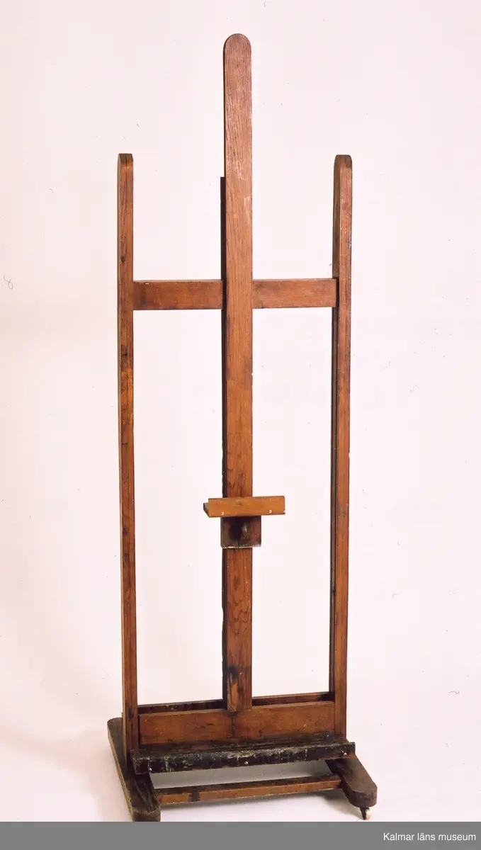 KLM 31949. Staffli. Tillverkat av ek. Staffliet kan användas från båda sidor. Staffliet består av tre parallella stolpar. På den mittersta stolpen löpes tavelstödet. Nedtill på båda sidorna finns en hylla för penslar, färg m.m. Staffliet går på fyra små mässingshjul.