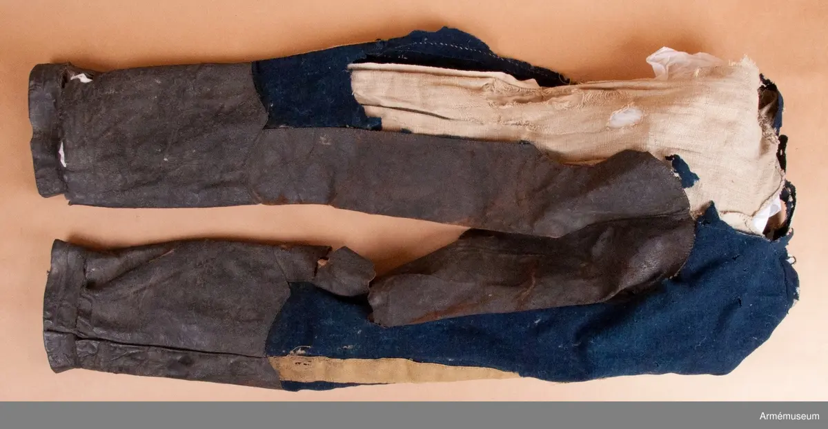 Blå ridbyxor med gula revärer och skinnskodda ben och byxbak.

Byxorna är sydda av ett ganska tunt blått kommisskläde, och är helfodrade med oblekt linne- eller hampalärft.
Modellen är konstruerad med två fram- och två bakstycken, och har hel lucka fram som har knäppts mot en gördel eller linning. Luckan har 5 knapphål fram, varav tre har knäppts mot gördeln. I luckans sidor upptill sitter två spänntampar för knäppning bak i ryggslutet. I sömmen bak sitter en infälld kil för reglering av midjevidden, samt en hank av läder för upphängning.
I sidan har byxorna revärar av 4 cm breda gula redgarnsband.
Byxbenen är skodda med brunt skinn i en höjd av ca 36 cm nertill. Kanten är formad i två bågar fram och en spets bak. Skinnskoningen fortsätter på baksidan upp på insidan av låren och i byxbaken.
Byxorna är mycket trasiga och malätna, och både yttertyg och foder har lagats ett flertal gånger under deras användningstid.
Samtliga knappar saknas förutom i höger byxben längst ner där det sitter rester av enkla skinnknappar i varje sida.

Märkt på insidan "LS No 2".