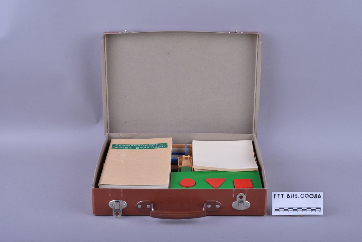Rektangulær brun koffert av papp, med håndtak og lås. Inne i kofferten er det små pappesker. Pappeskene inneholder små treklosser, plater med pålimte miniatyr-leker. Blant annet dukke, hund seng, saks, pipe. I tillegg er det flere små knapper, notatblokk og tekstilbånd. I kofferten ligger også en bok merket "TERMAN-MERRILL // NORSK STANDARD".